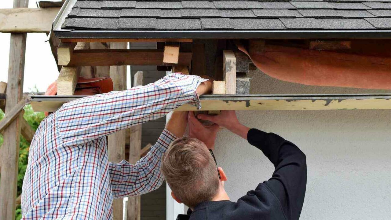 two men father and teen son installing wooden plank frame for eaves of gray asphalt shingles roof